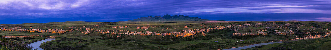 A panorama of the sandstone landscape in blue-hour twilight at Writing-on-Stone Provincial Park (Áísínai'pi) in Alberta, with the Milk River below winding amid the sandstone rock formations, and the Sweetgrass Hills in the distance in Montana.