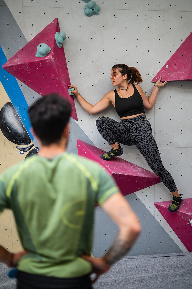 Young man teaching woman in her twenties how to climb on a climbing wall indoors