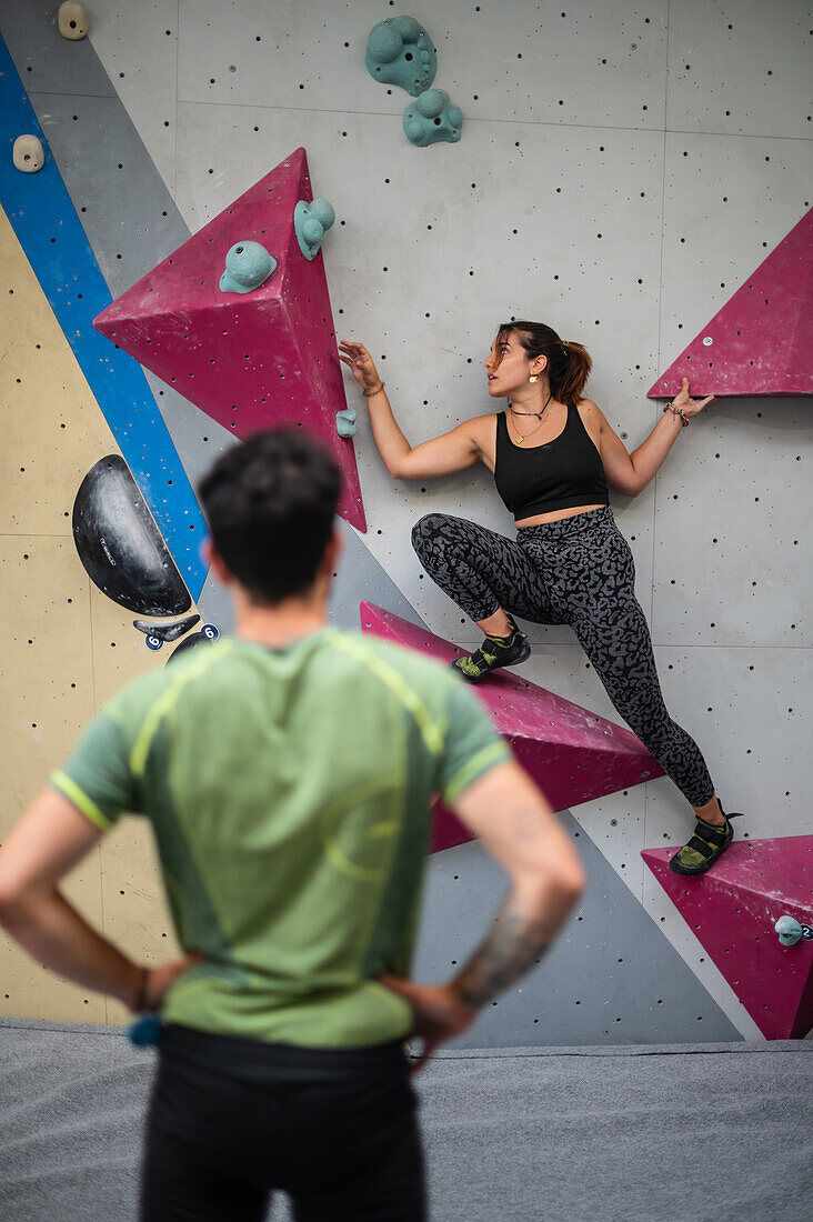 Young man teaching woman in her twenties how to climb on a climbing wall indoors