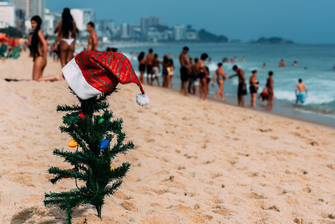 Ein behelfsmäßiger kleiner traditioneller Weihnachtsbaum und eine Weihnachtsmannmütze, die im Sand am Leblon Beach aufgestellt sind, mit einer unscharfen Menschenmenge im Hintergrund, Rio de Janeiro, Brasilien, Südamerika