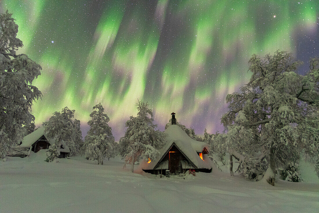 Winteransicht einer typischen Holzhütte, die vom Feuer im gefrorenen Wald während eines Nordlichtsturms beleuchtet wird, Finnisch-Lappland, Finnland, Europa