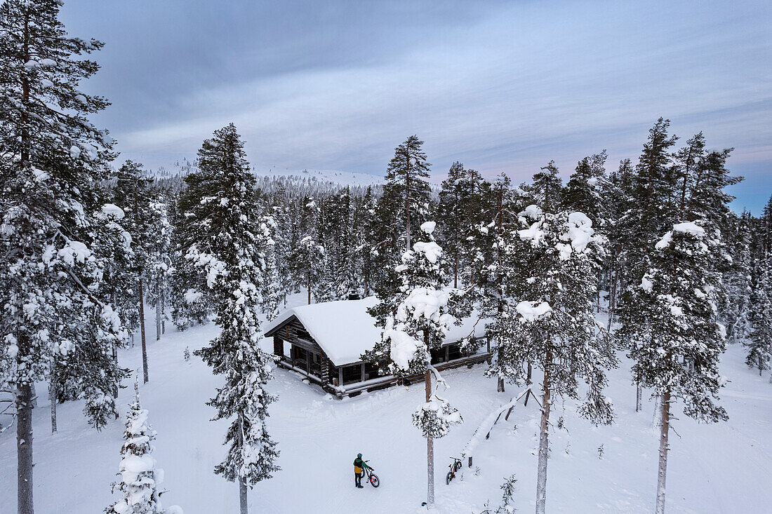 Drohnenaufnahme eines Radfahrers mit Fatbike, der inmitten eines verschneiten Nadelwaldes vor einer Berghütte steht, Finnisch-Lappland, Finnland, Skandinavien, Europa