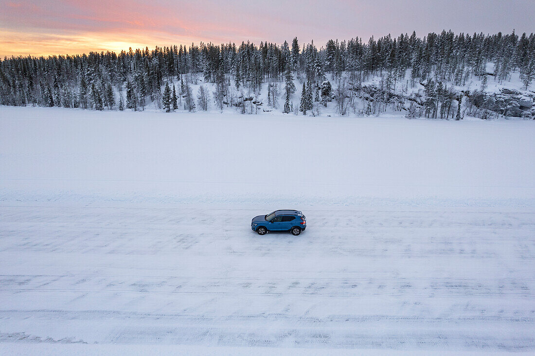 Drohnenaufnahme eines blauen Geländewagens, der in der Mitte eines leeren, zugefrorenen Sees in der Winterlandschaft des Polarkreises steht, Jokkmokk, Norrbotten, Schwedisch-Lappland, Schweden, Skandinavien, Europa