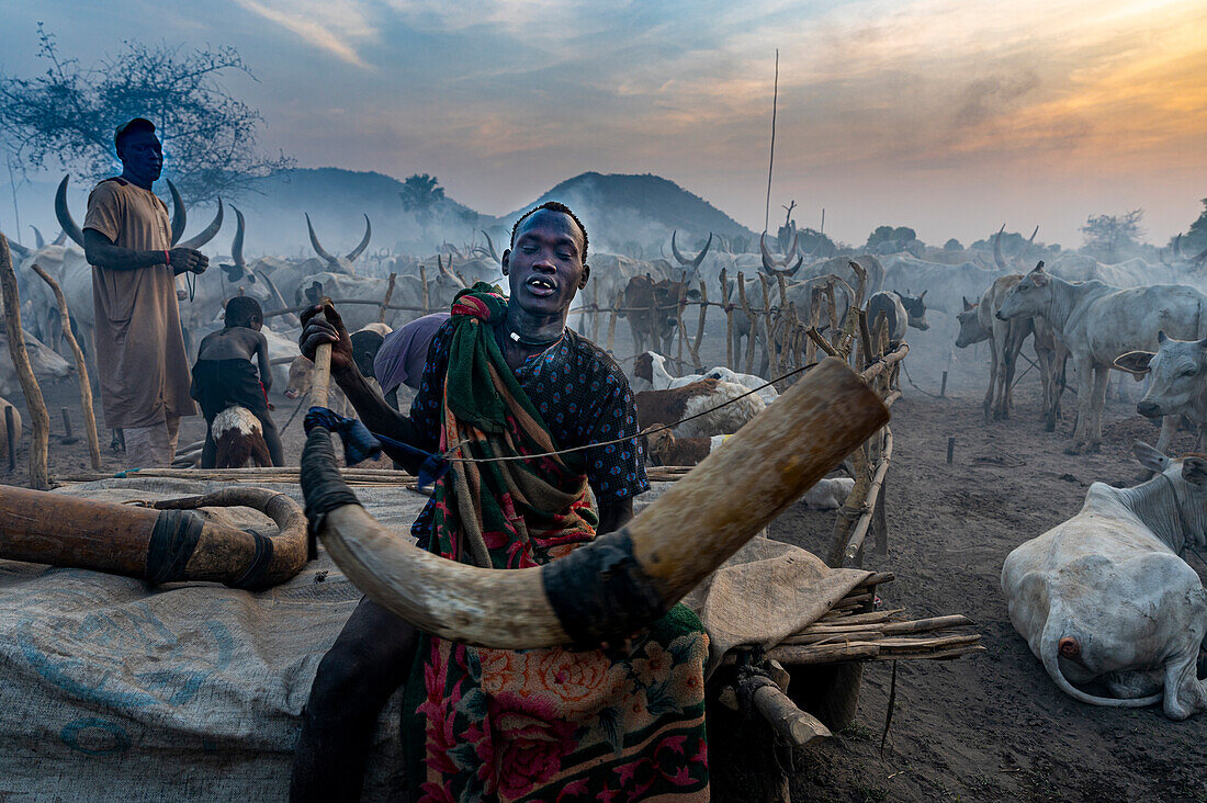 Mann bläst in ein massives Kuhhorn, um den Kühen zu signalisieren, dass sie zurückkommen sollen, Mundari-Stamm, Südsudan, Afrika