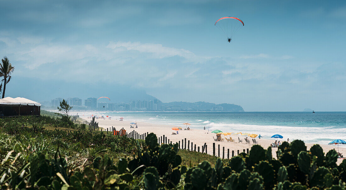 Ein Gleitschirmflieger schwebt über einem ruhigen, mit bunten Sonnenschirmen übersäten Strand mit einem klaren blauen Himmel im Hintergrund in Barra da Tijuca, Rio de Janeiro, Brasilien, Südamerika