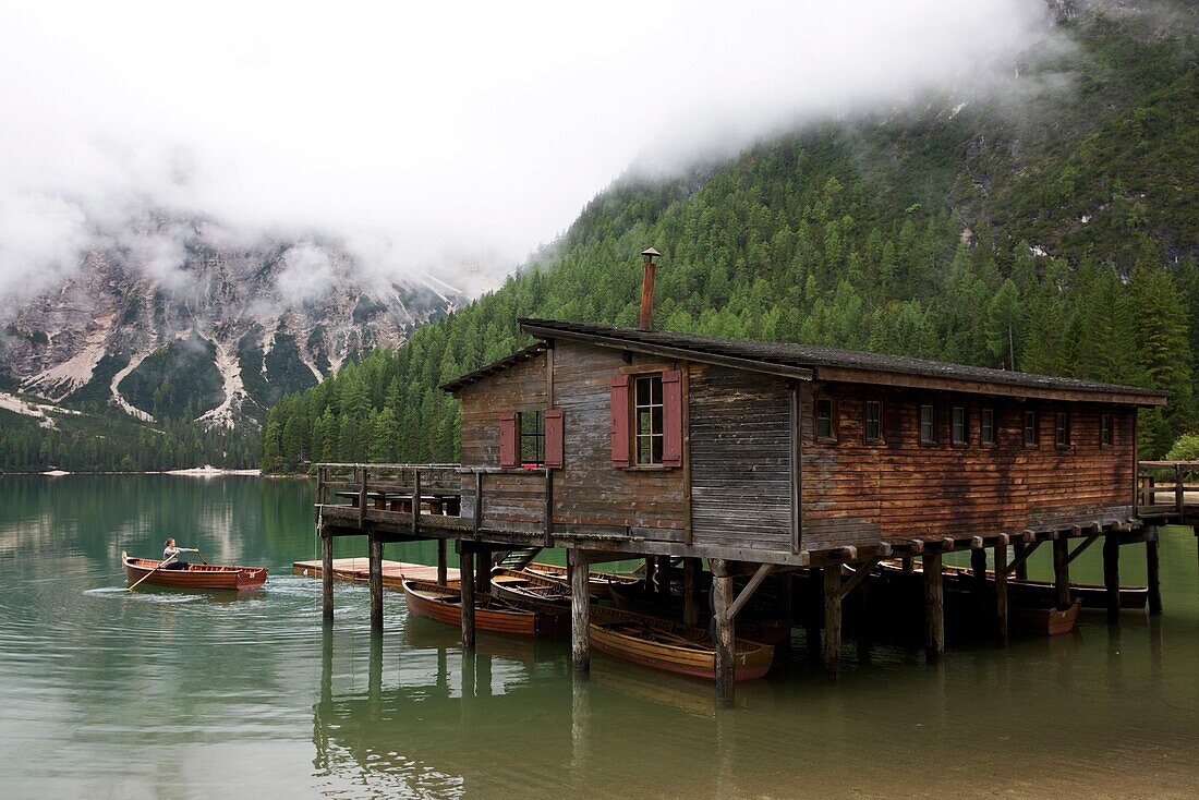 Italien, Autonome Provinz Bozen, Hochpustertal, Pragser Wildsee, Frau rudert in einem Boot vor einem Holzhaus auf Stelzen auf dem smaragdgrünen Wasser des Sees, umgeben von grünen Bergen im Nebel