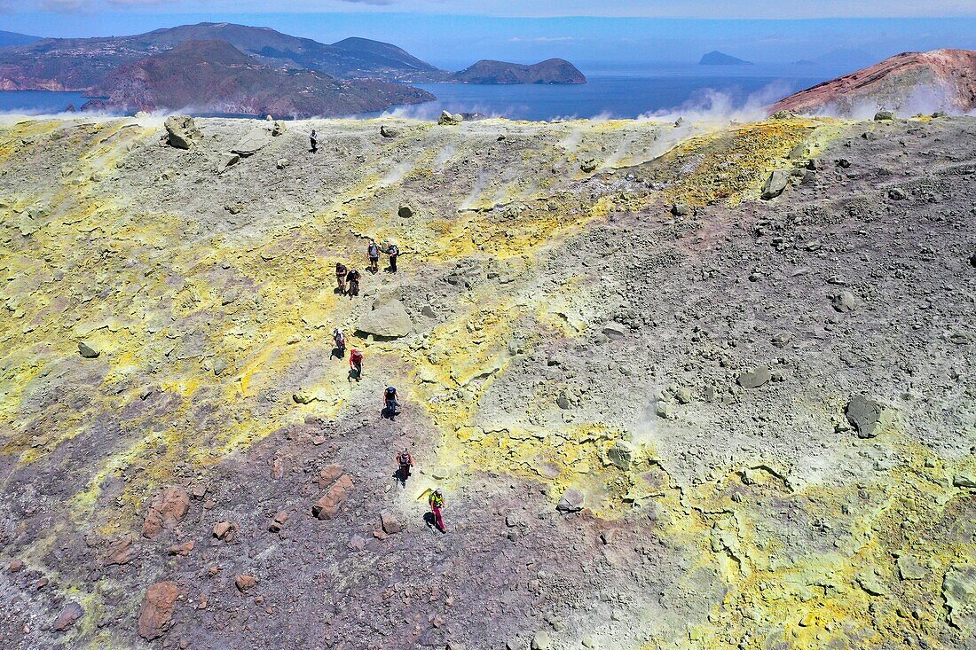 Italien, Sizilien, Äolische Inseln, UNESCO-Welterbe, Insel Vulcano, Wanderer am Kraterrand des Vulkans della Fossa beim Gang durch Schwefelfumarolen, links im Hintergrund die Insel Lipari und rechts Panarea und Stromboli (Luftaufnahme)