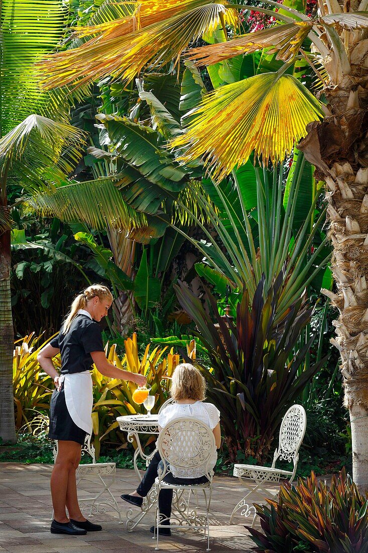 Spanien, Kanarische Inseln, La Palma, Kellnerin serviert einem Kunden ein Glas Orangensaft in den üppigen tropischen Gärten eines Luxushotels