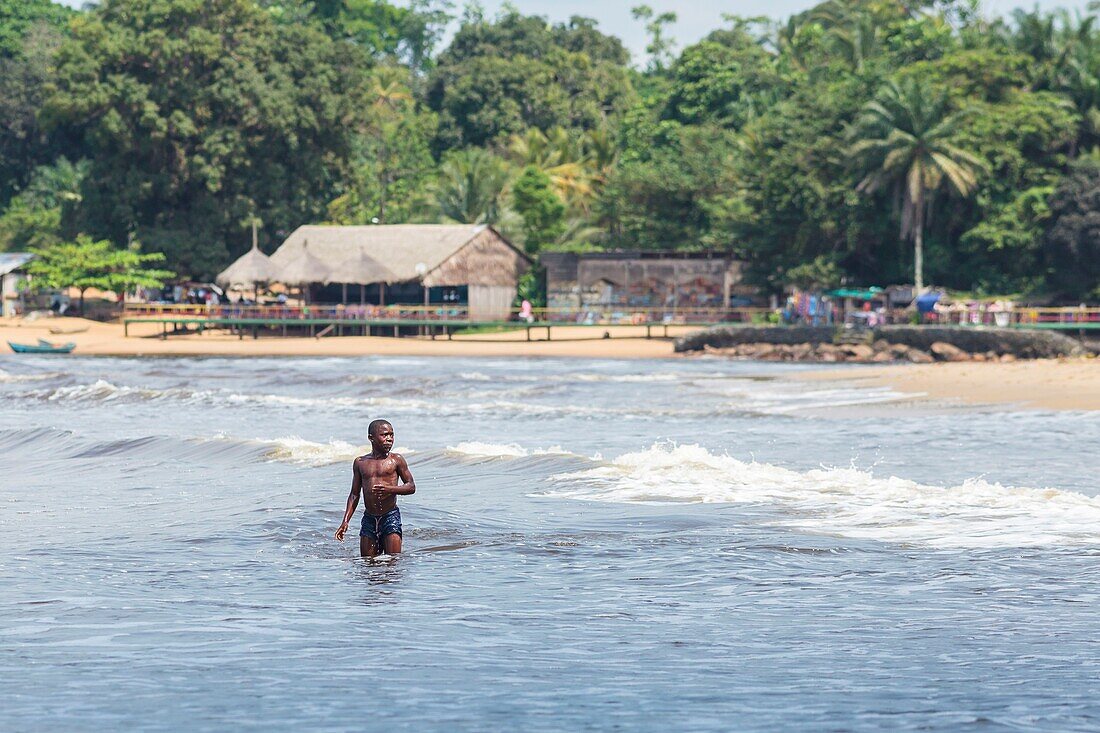 Cameroon, South Region, Ocean Department, Kribi, African boy playing in waves in front of a fishing village