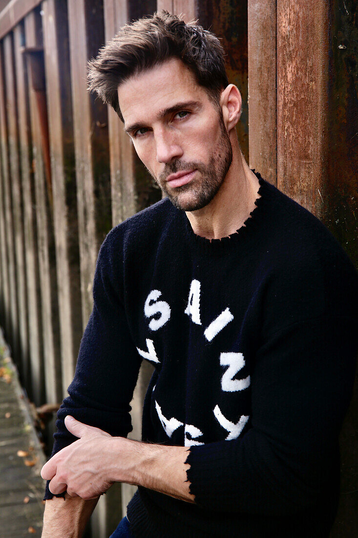 Man in black knitted sweater with white lettering in front of wooden picket fence