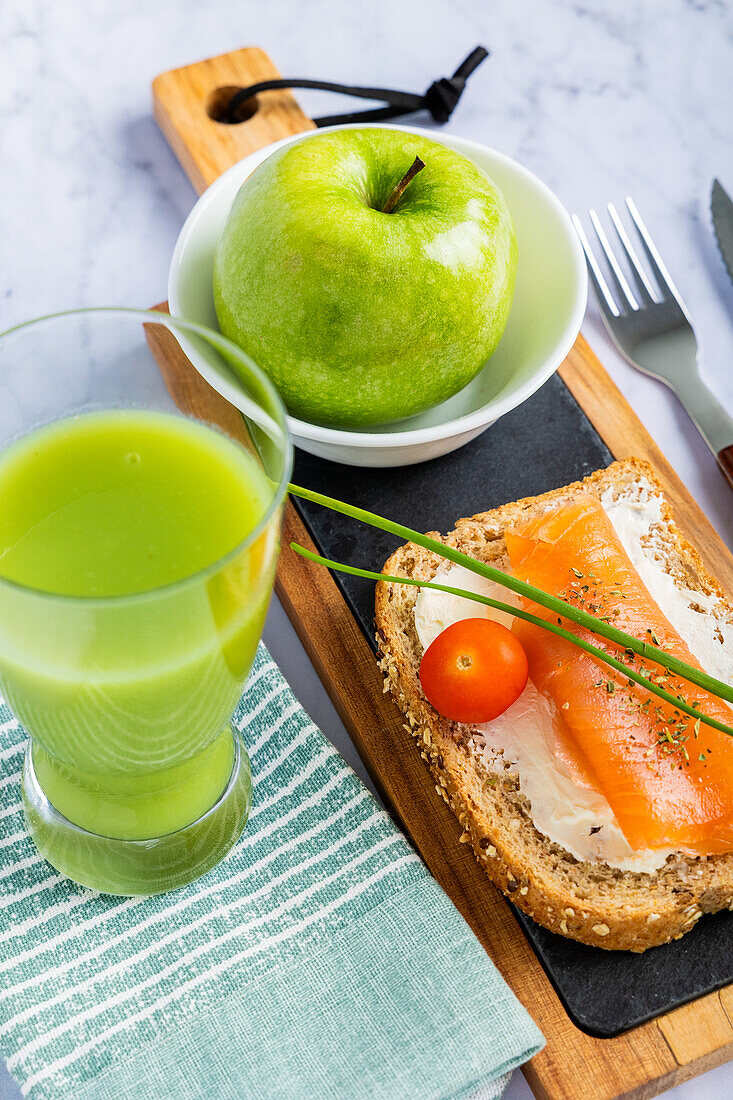 Blick von oben auf eine nahrhafte Mahlzeit mit einer Scheibe Vollkorntoast, belegt mit Räucherlachs, Frischkäse und einer Kirschtomate, dazu ein frischer grüner Apfel-Gurkensaft. Serviert auf einem Holzbrett mit einem modernen Marmorhintergrund.