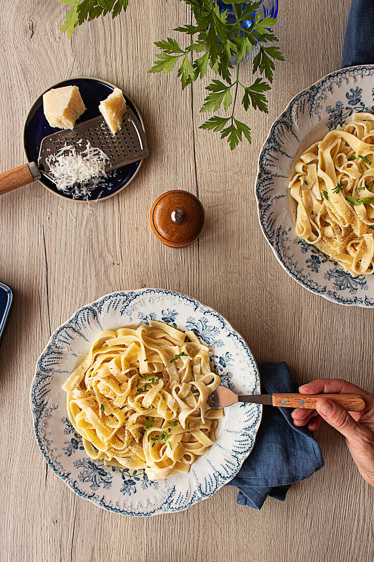 Nudelgericht Alfredo mit cremiger weißer Soße und Kräutern, serviert auf einem Tisch mit Pfeffermühle, Parmesanreibe und blauer Serviette (von oben)