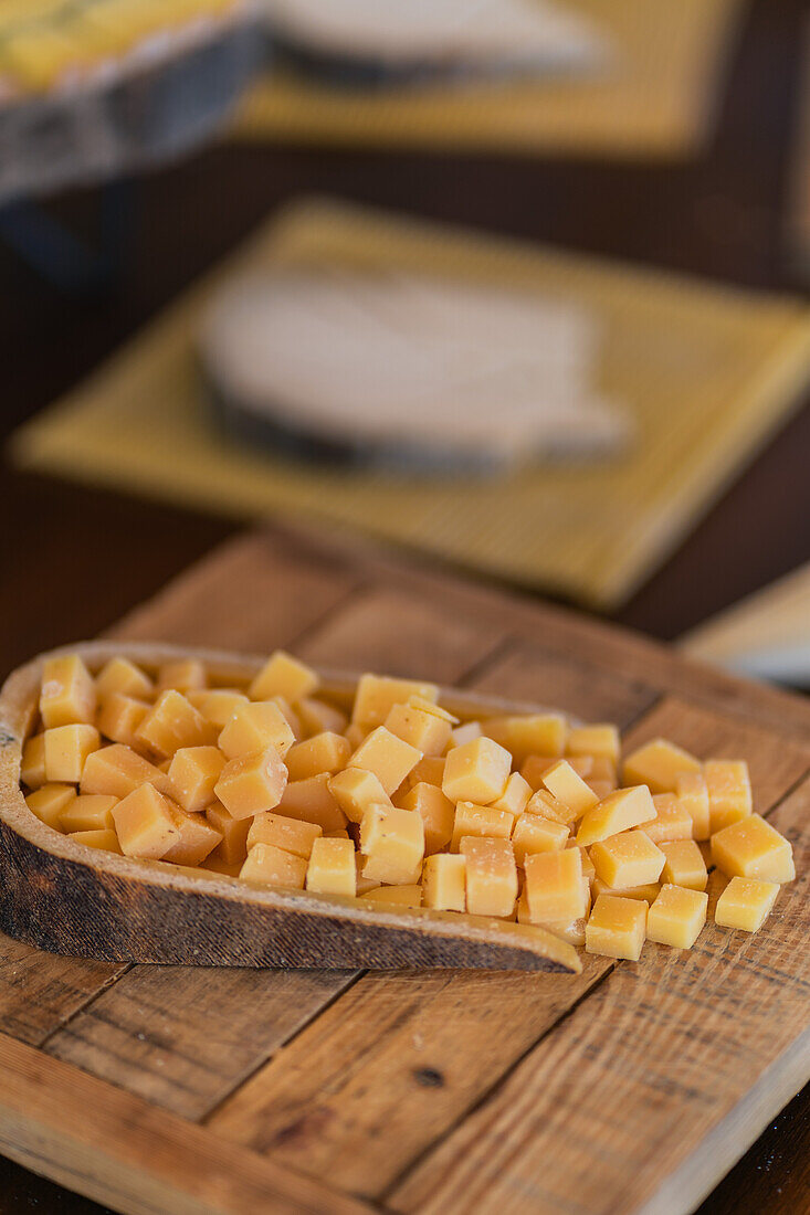 Gewürfelter, handwerklich hergestellter Gouda-Käse auf einem geschnitzten hölzernen Servierbrett, mit Weichzeichner-Käse im Hintergrund