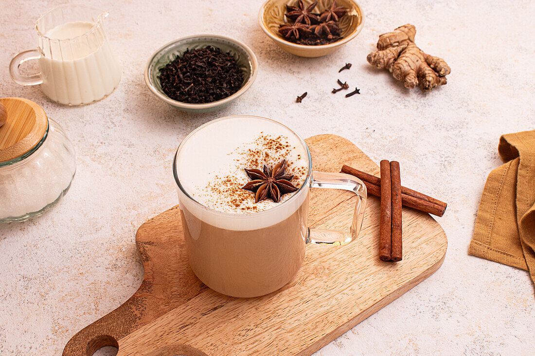 Von oben, köstlicher Chai Latte mit cremigem Schaum auf einem Holzbrett, umgeben von Sternanis, Zimtstangen, frischem Ingwer, Teeblättern, einem Milchglas und einer Serviette