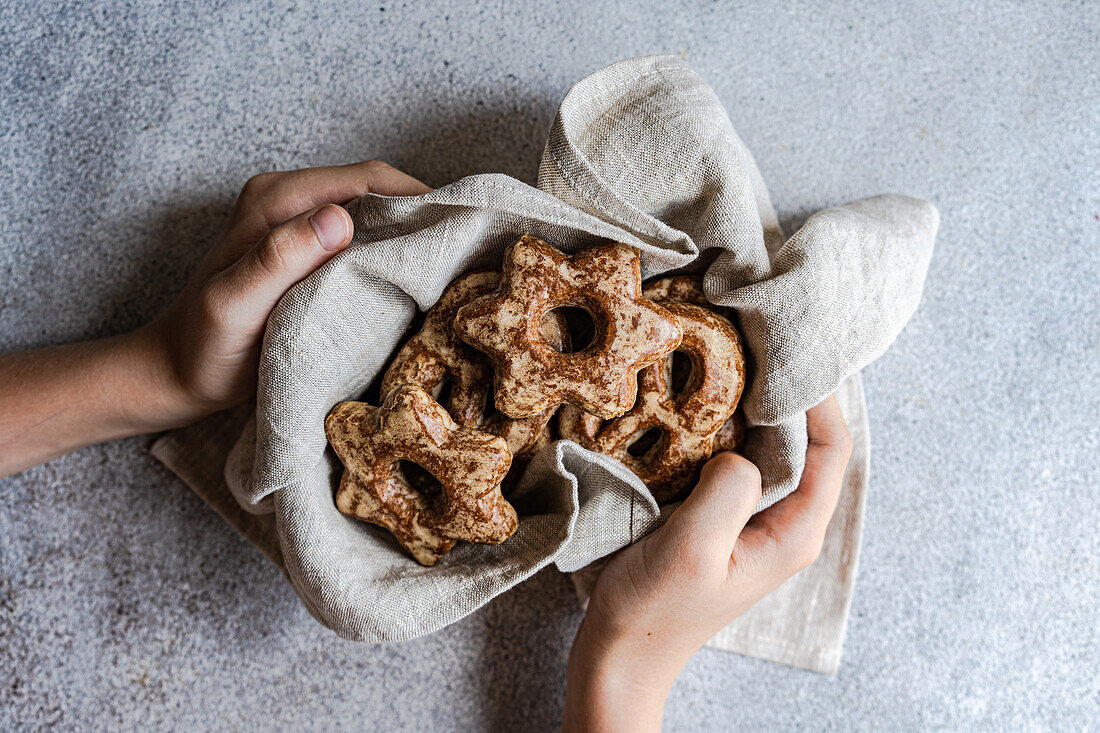 Draufsicht auf abgeschnittene, nicht erkennbare Hände, die ein Tuch halten, das mit selbstgebackenen Lebkuchen in Form von Sternen gefüllt ist, die auf einer strukturierten grauen Oberfläche präsentiert werden.