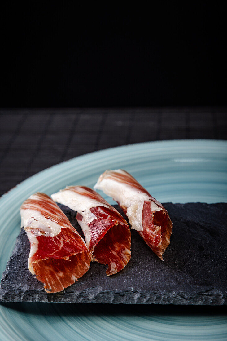 Serrano-Schinken, eine begehrte spanische Delikatesse, elegant präsentiert auf einer schwarzen Schiefertafel vor dunklem Hintergrund, der seine reiche Textur und Farben hervorhebt