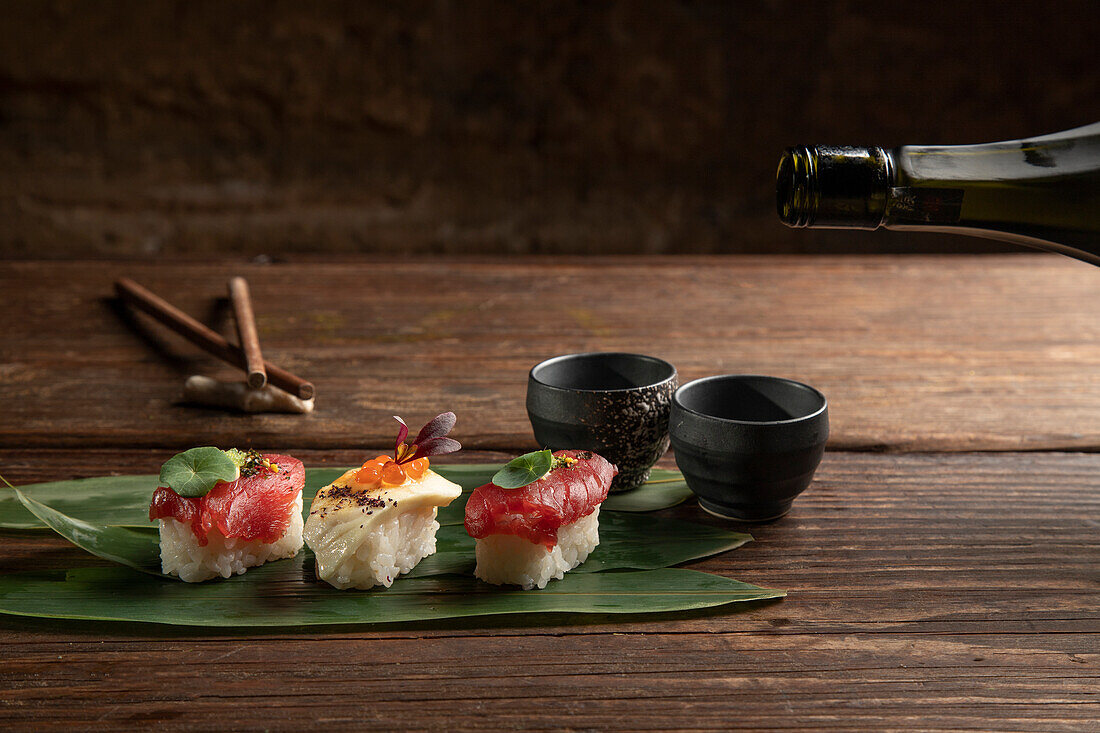 An elegant presentation of sushi delicacies, laid on a bamboo leaf, accompanied by sake cups, on a rustic wooden table setting, ideal for a sophisticated dining experience