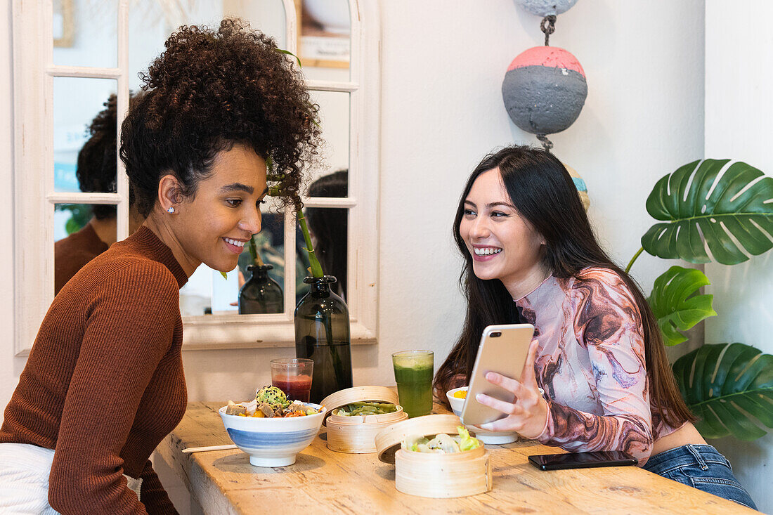 Fröhliche verschiedene weibliche Freunde sitzen am Tisch mit Poke im Restaurant und verwenden Smartphone, während sie das Wochenende zusammen verbringen