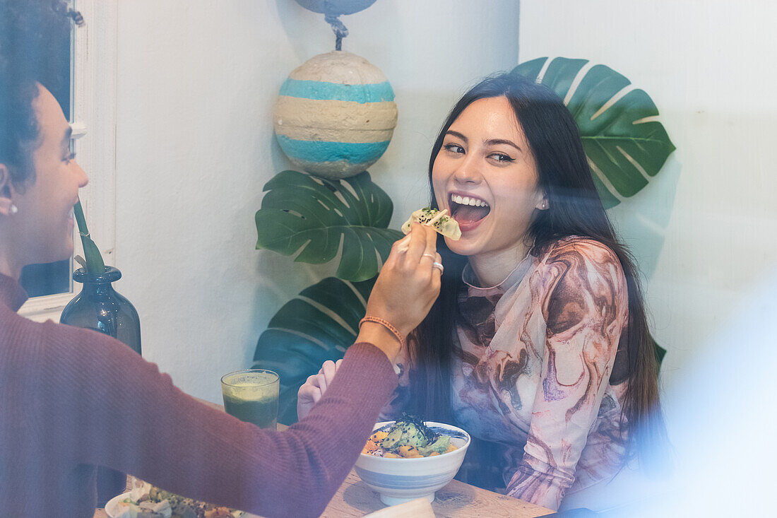 Through glass cheerful black woman feeding female friend with poke while chilling in restaurant together