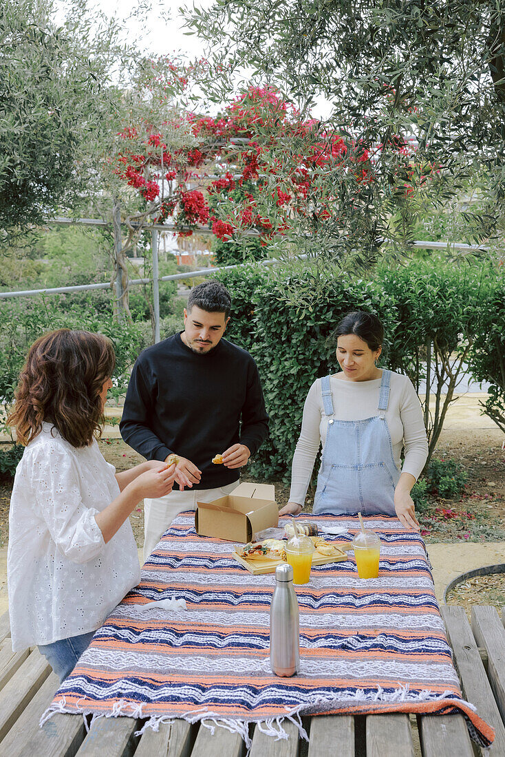 A diverse group of three friends, two women and one man, enjoys a lively picnic in a vibrant outdoor setting. The group engages in conversation while sharing food from a single box on a colorful striped blanket, under flowering trees