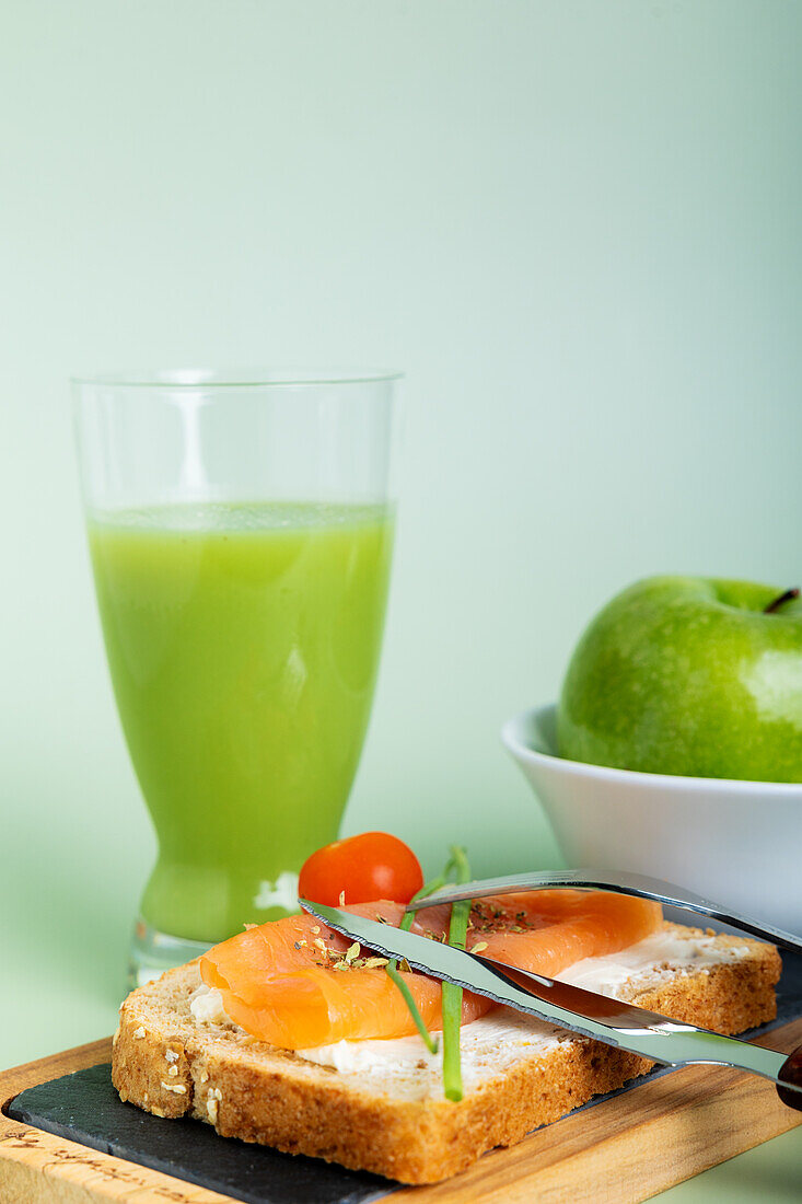 In a vibrant display, this image captures a wholesome breakfast spread featuring a glass of fresh cucumber and apple juice, a salmon sandwich on multi-grain bread adorned with cherry tomatoes and chives, alongside crisp green apples.