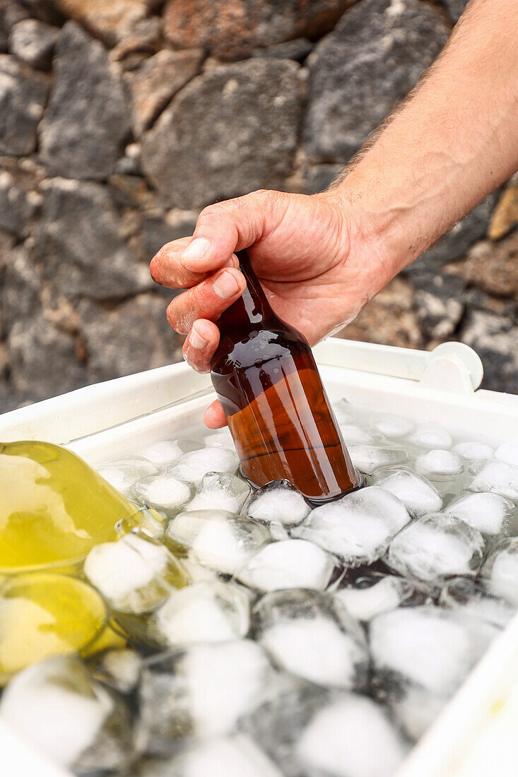 Abgeschnittene, nicht erkennbare Männerhand, die eine gekühlte braune Bierflasche aus einem mit Eis gefüllten Kühler ergreift, vor einem steinernen Hintergrund im Freien.