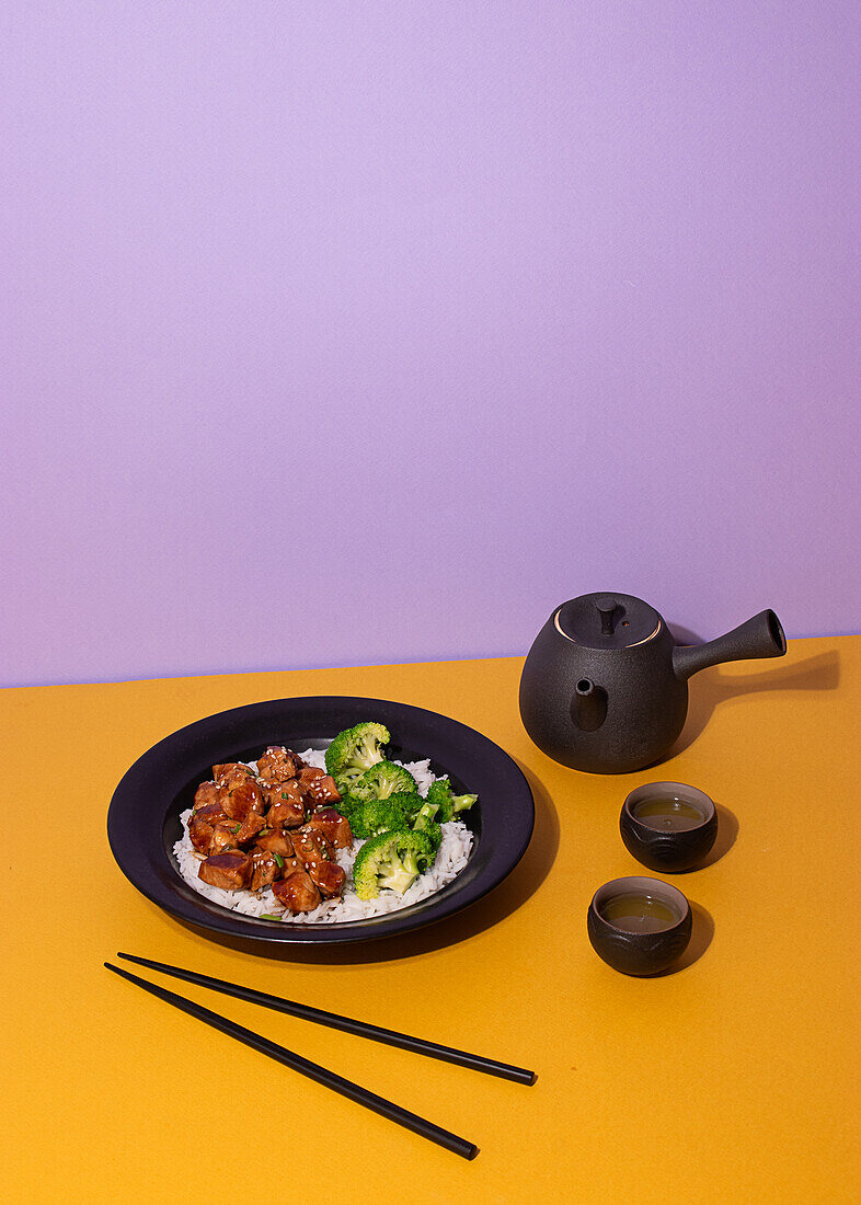 Kunstvoll angerichtetes Teriyaki-Hähnchen auf einem schwarzen Teller, serviert mit Brokkoli und Reis, begleitet von einem traditionellen Teeservice auf einem leuchtend gelben und violetten Hintergrund.