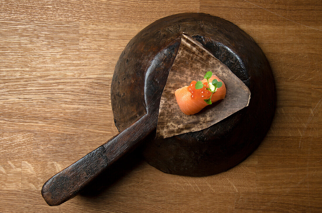 A single piece of expertly cut salmon sushi, elegantly presented in a rustic, aged container Small green leaves and red eggs add freshness and color to the composition