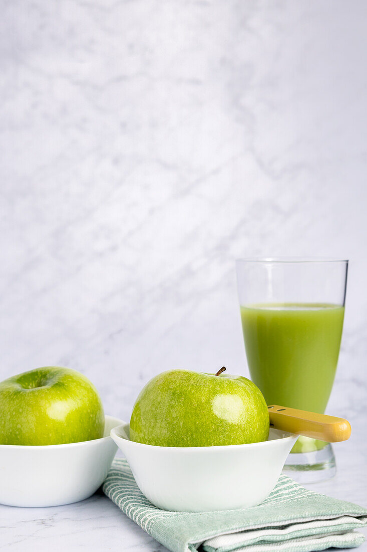 Frische grüne Äpfel in einer weißen Schale neben einem Glas mit nahrhaftem Gurken- und Apfelsaft, elegant auf einer Marmorfläche platziert. Die Szene wird durch eine zartgrüne Serviette ergänzt, die einen gesunden Lebensstil unterstreicht.