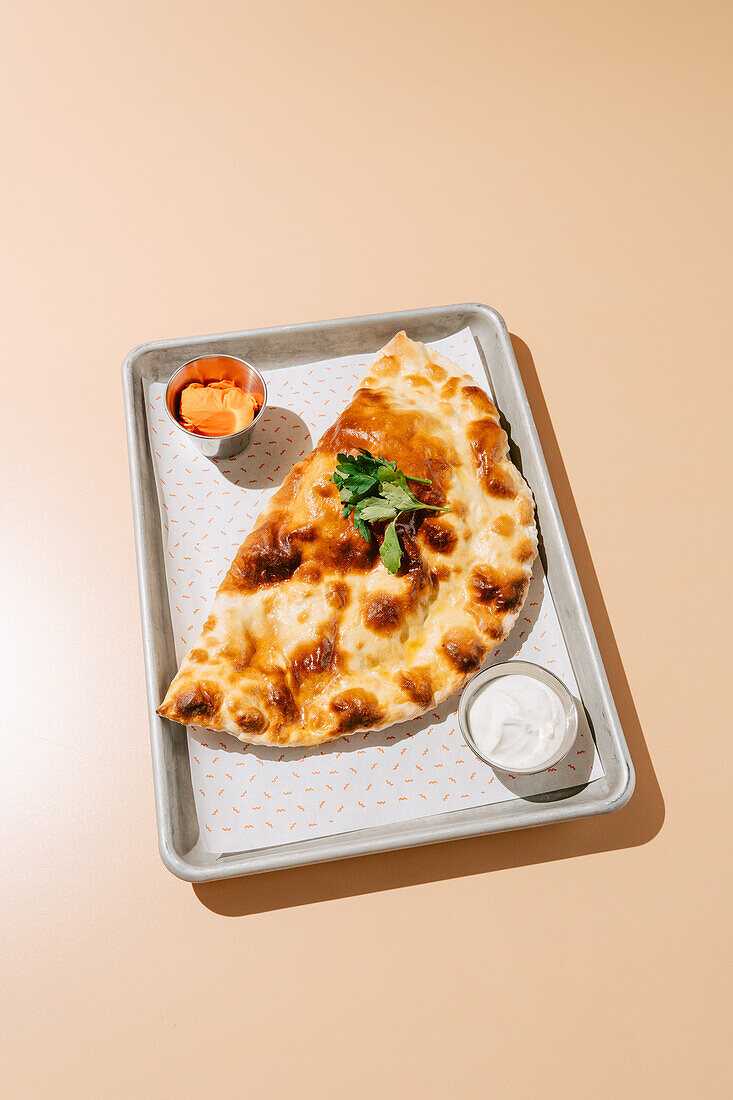 From above golden-brown baked cheburek paired with a sour cream-garlic sauce and a spicy hot sauce, served on a minimalist tray with a garnish of fresh parsley.