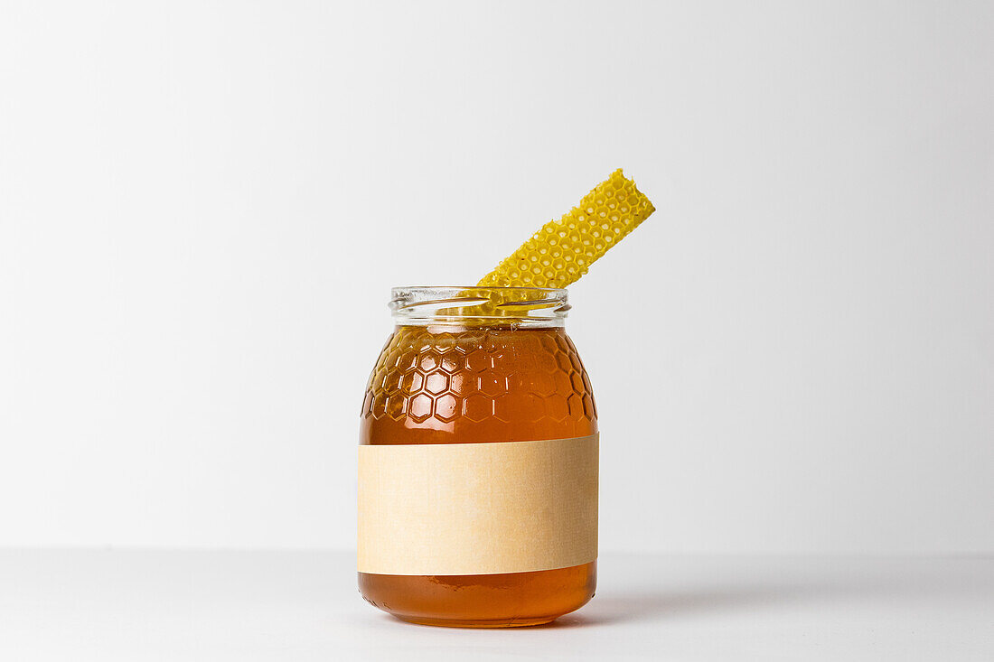 Piece of honeycomb on top of glass jar full of fresh natural golden honey with white background