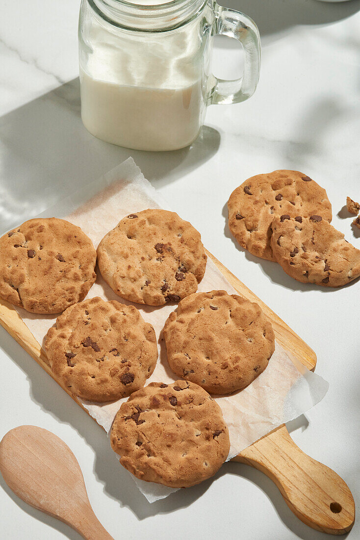 Leckere Schokokekse auf einem Backpapier neben einem Glas Milch und einem Holzlöffel, aufgenommen von oben bei natürlichem Licht