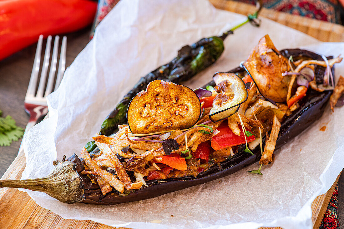 Baked stuffed eggplant with cherry tomatoes, bell peppers, onion, garlic, thin potato slices, cheese, and microgreens served on parchment paper."