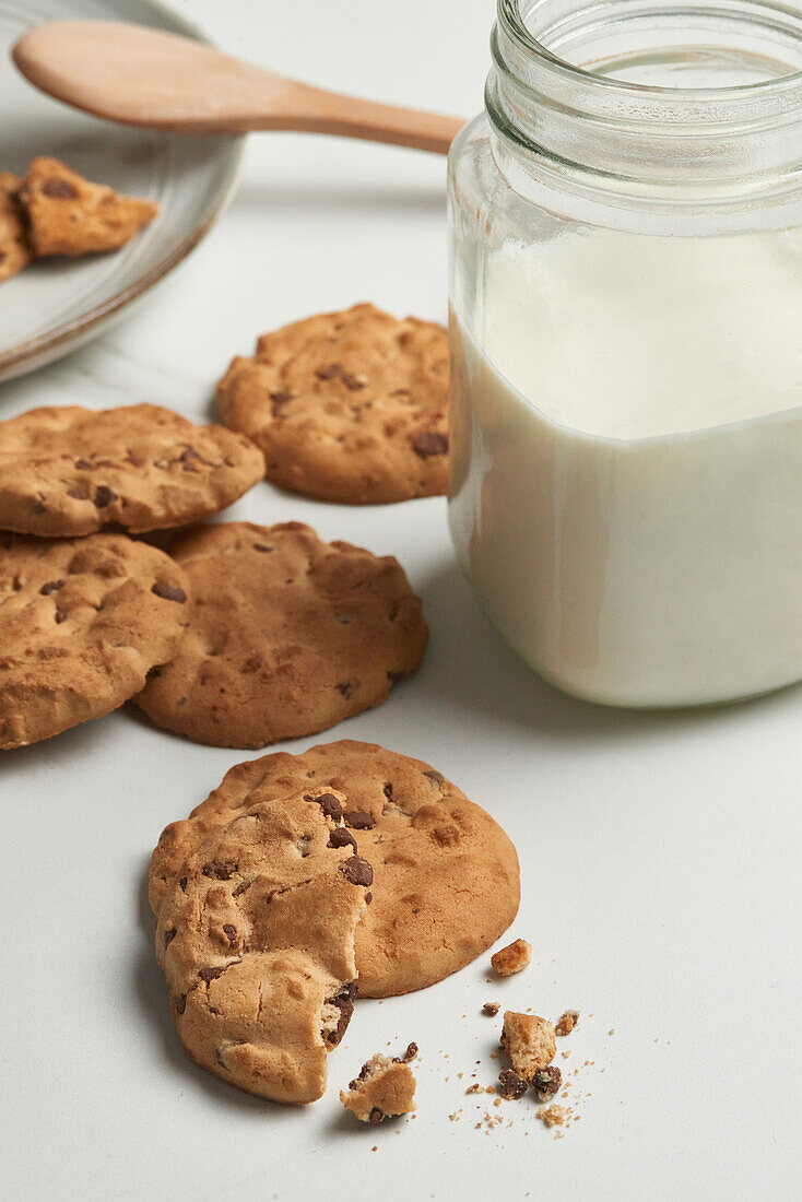 A top-down view of a homely snack scene with chocolate chip cookies and a jar of milk on a white surface, embodying a comforting treat