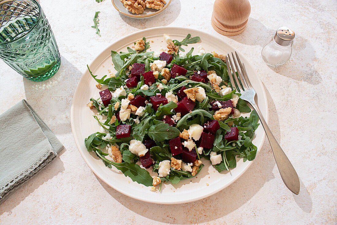 Healthy goat cheese, arugula and beetroot salad on a beige plate and styled with a pepper mill, a lime water glass on a rustic but vibrant atmosphere