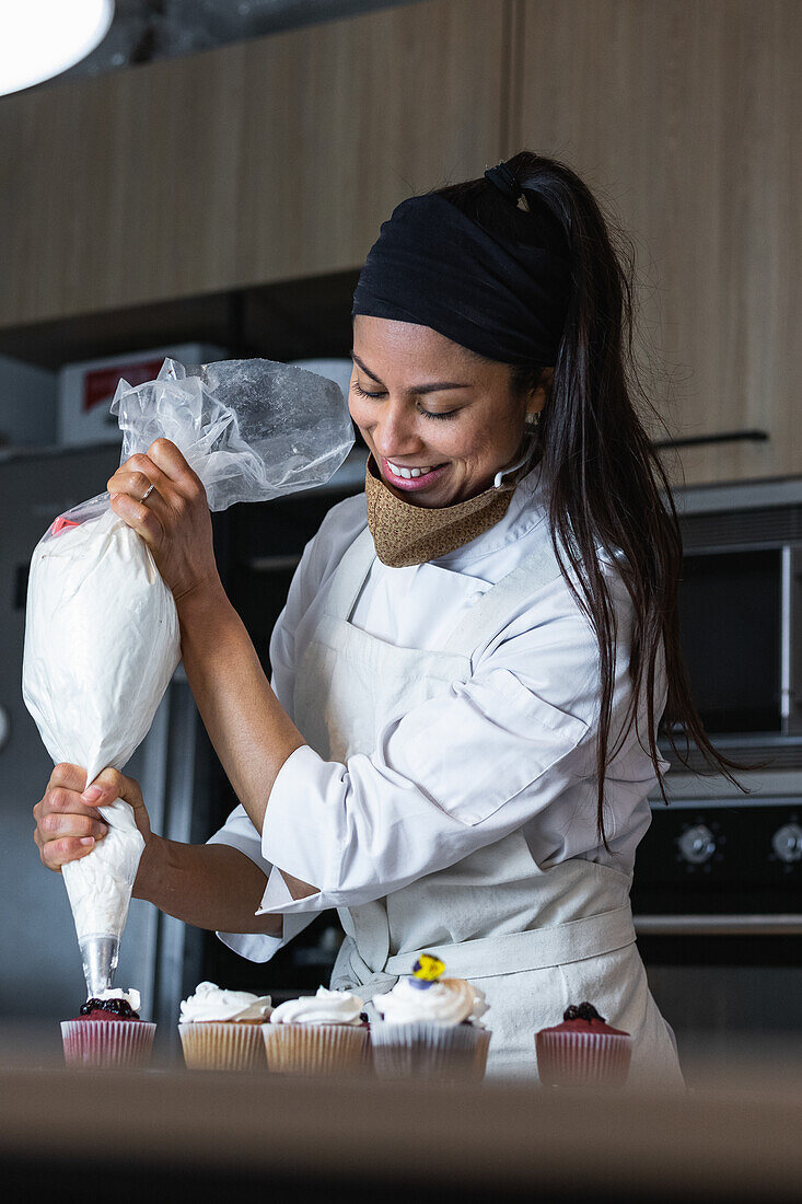 Fröhliche Bäckerin, die beim Backen von Desserts in einer Backstube Schlagsahne aus einem Spritzbeutel auf leckere Cupcakes spritzt