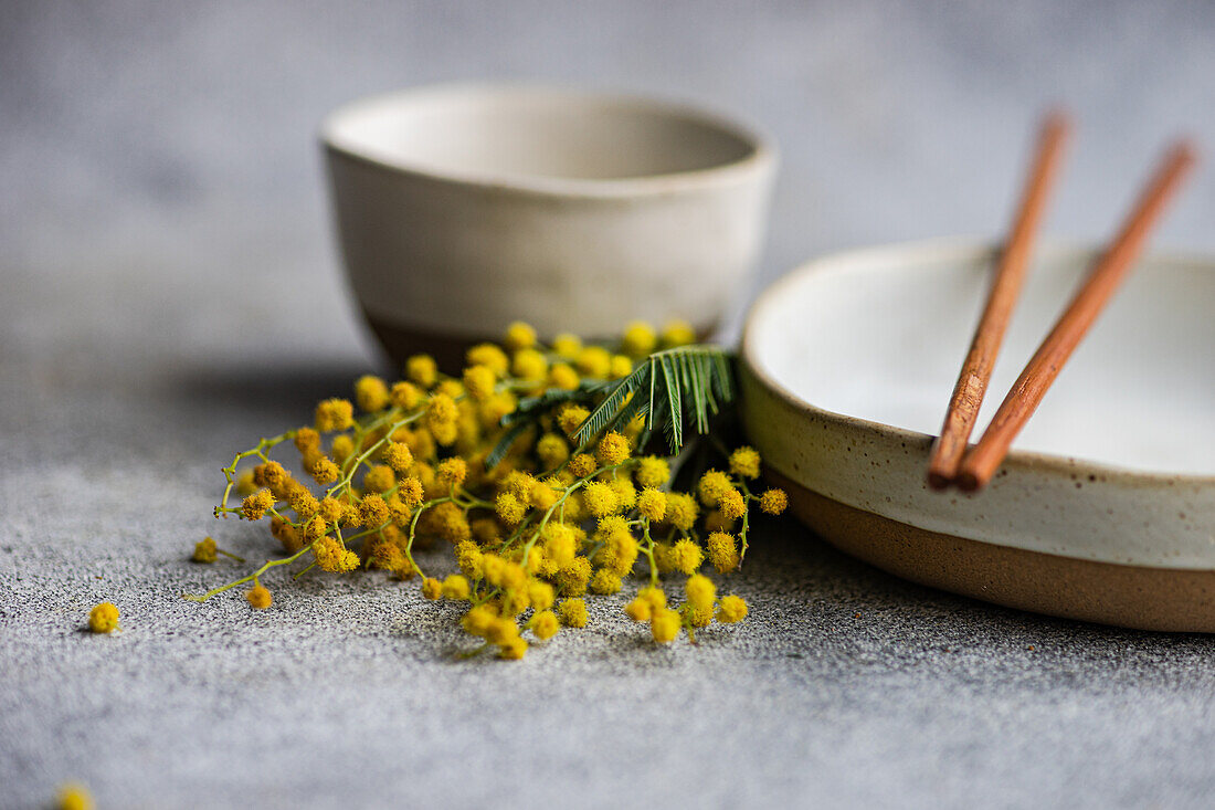 Ein ruhig gedeckter Tisch mit Keramikgeschirr, Kupferstäbchen und leuchtenden Mimosenblüten als Farbakzent