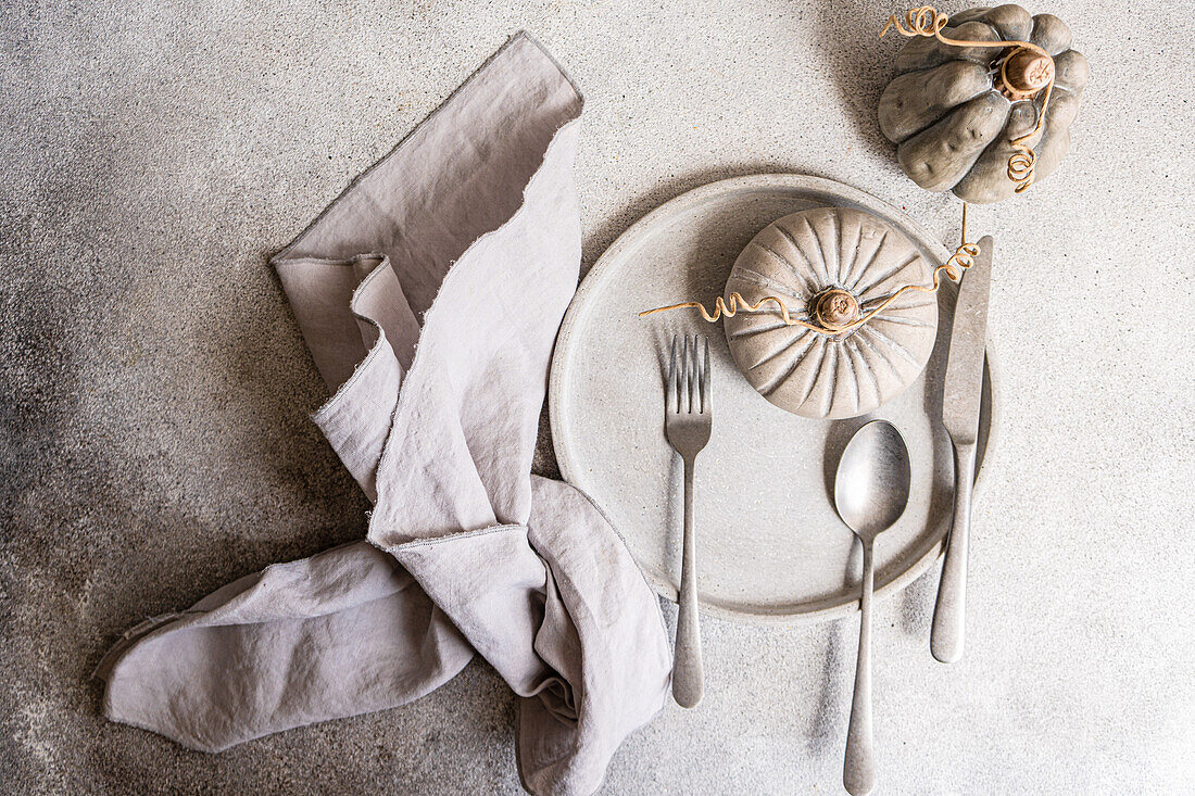 Ein geschmackvolles, herbstliches Flatlay mit einem neutral gedeckten Tisch mit einem Zierkürbis, grauen Tellern und einer Leinenserviette - ideal für saisonale Designthemen