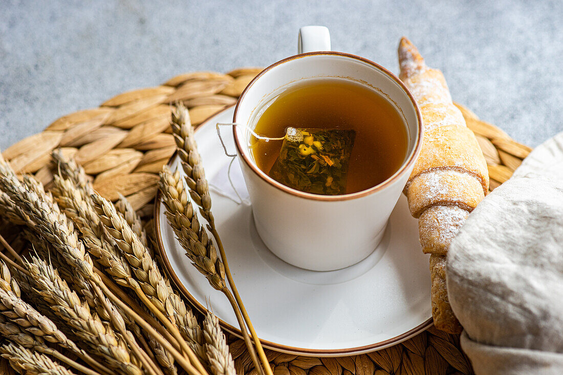 Eine warme Tasse Schwarztee mit einem Teebeutel, der mit getrockneten Zitronenstücken durchzogen ist, begleitet von einem frischen, hausgemachten Croissant, das leicht mit Puderzucker bestäubt ist und elegant neben Weizenähren auf einem gewebten Tischset präsentiert wird.