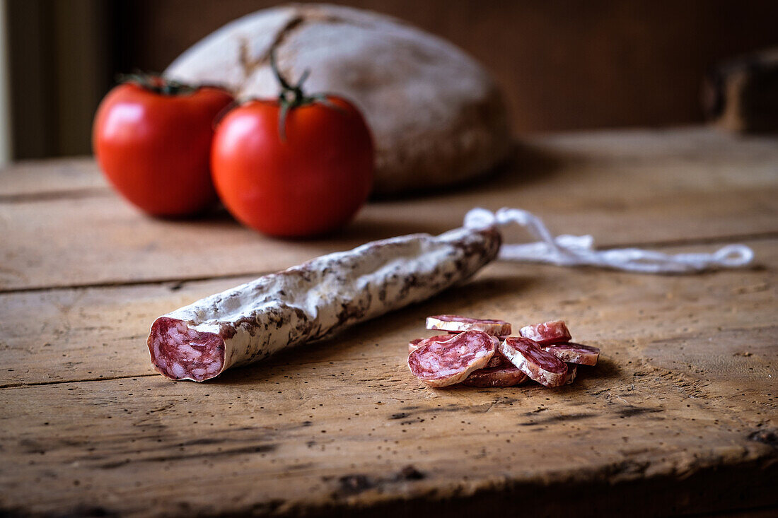 A rustic composition featuring a sliced cured fuet, alongside fresh tomatoes and a loaf of bread, all set on a well-worn wooden table.