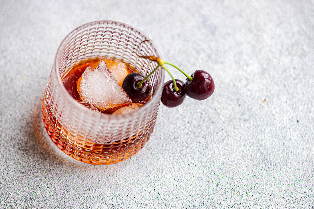 A sophisticated glass of cherry liqueur served on ice, garnished with a pair of ripe cherries, presented on a textured surface