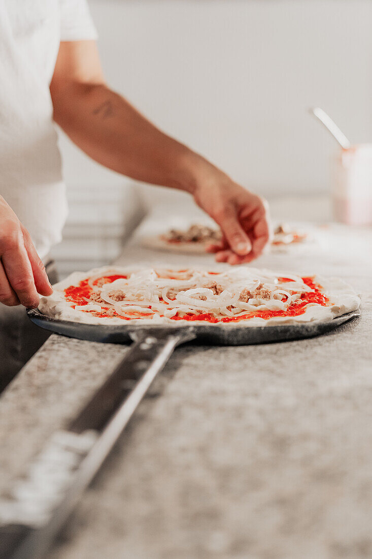 An anonymous chef in a pizzeria skillfully adds toppings to a fresh pizza on a peel, in a faceless crop focusing on the hands