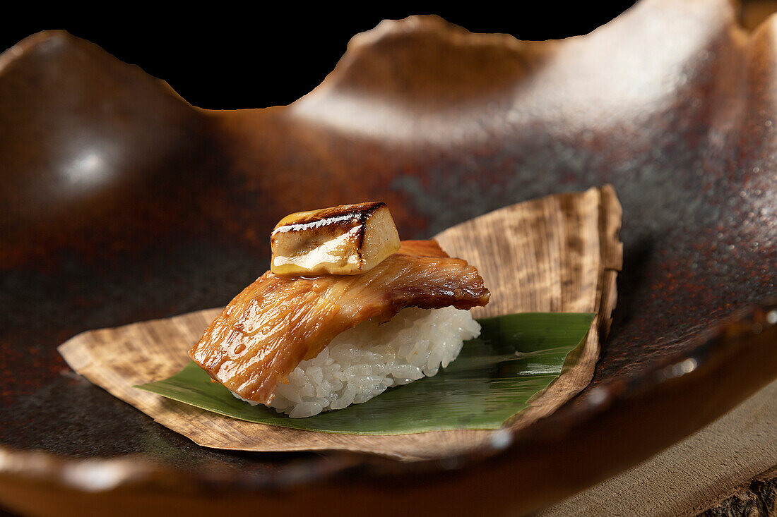 A beautifully crafted sushi piece, featuring grilled fish atop a bed of sticky rice, elegantly presented on a bamboo leaf and rustic ceramic dish