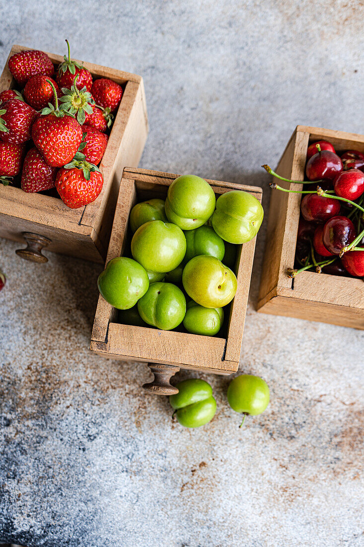 Wooden boxes of organic ripe berries—green plums, cherries, and strawberries—on a textured backdrop