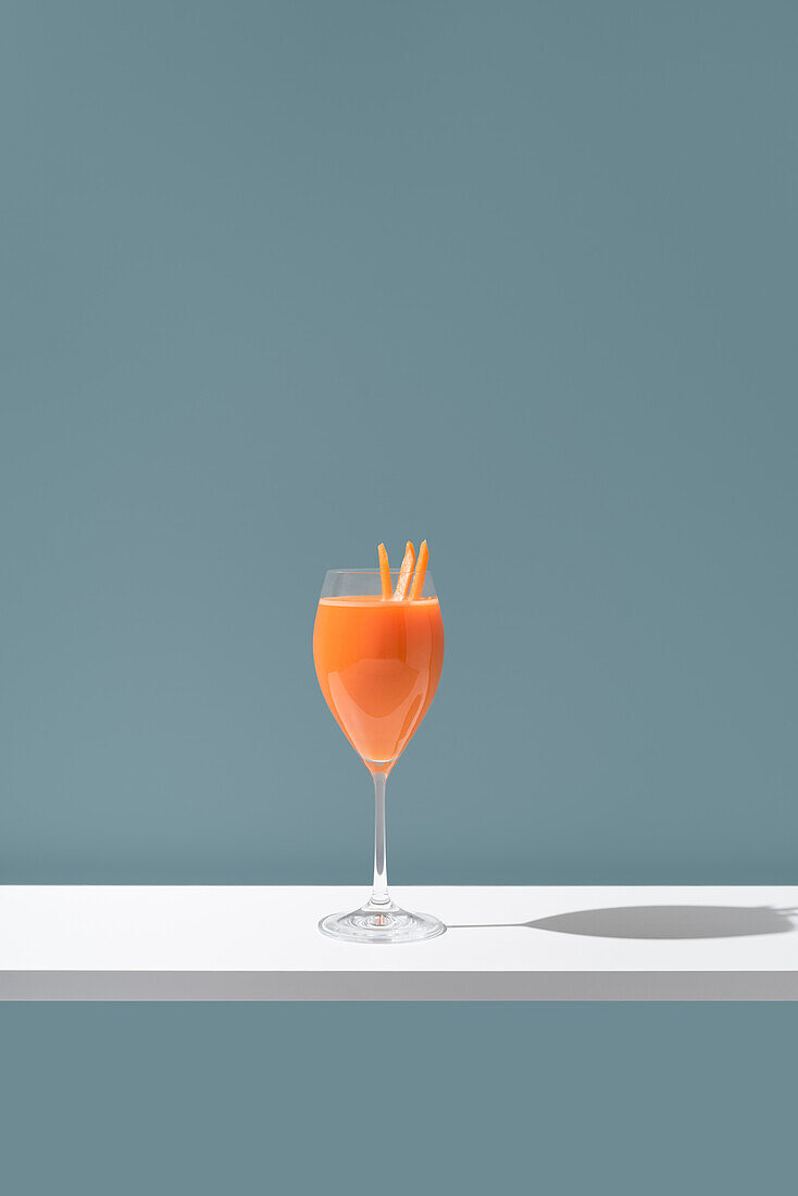 A minimalistic image showcasing an orange-colored beverage elegantly presented in a wine glass, adorned with orange slices as garnish against a soft blue background.