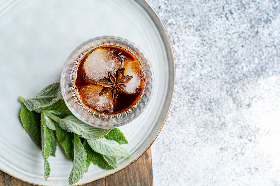 Blick von oben auf einen erfrischenden Kirschcocktail mit Eis und einem Zweig Minze, präsentiert auf einem rustikalen Teller mit einer Sternanisgewürz-Garnitur