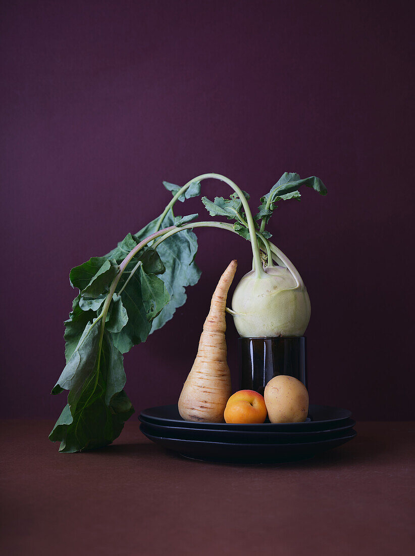 A creative still life composition of root vegetables including a parsnip, kohlrabi with leaves, and small potatoes, elegantly arranged on a dark, reflective plate against a deep maroon backdrop