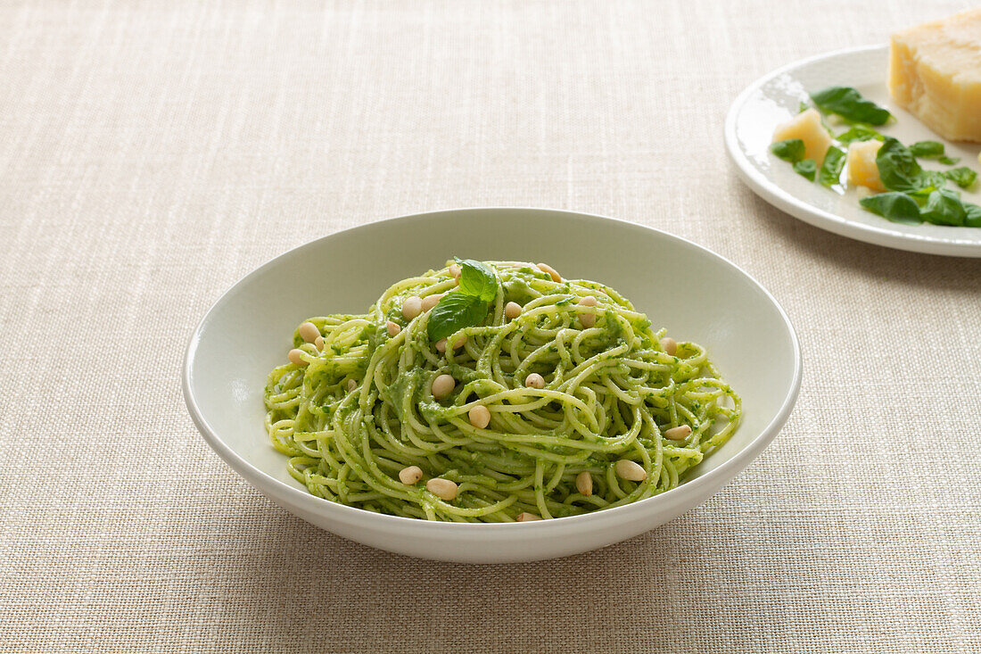 A bowl of homemade spaghetti pesto garnished with fresh basil leaves and pine nuts on a textured tablecloth.