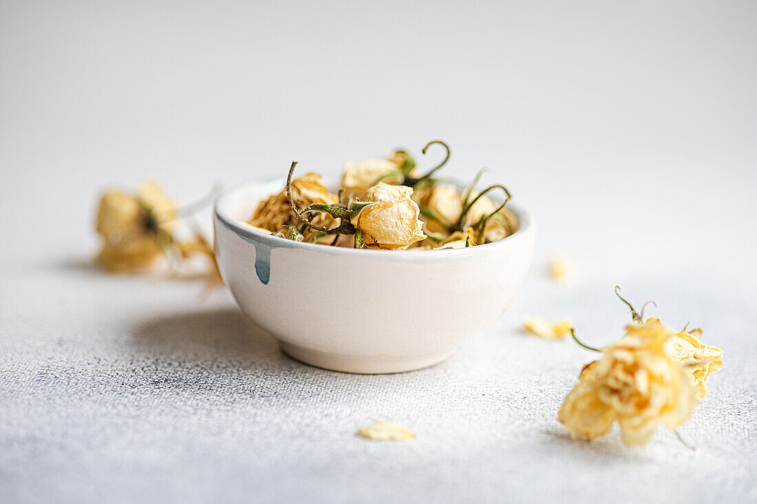 A ceramic bowl filled with delicate dried roses rests against a soft, textured white background, offering a simplistic yet elegant decorative theme