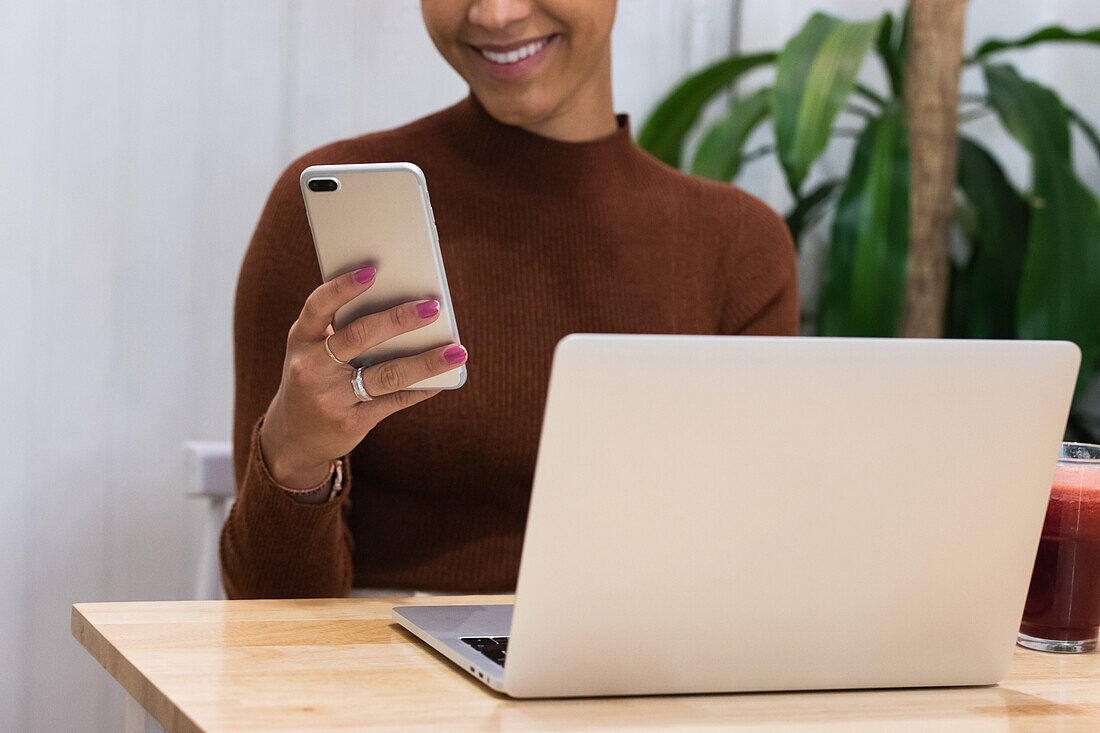 Cropped unrecognizable happy African American female freelancer browsing smartphone and smiling while sitting at table with laptop in cafe and working on remote project